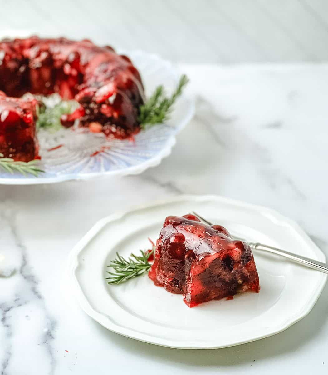 Slice of cranberry jello salad mold on a small white plate with a fork and rosemary, with the rest of the mold on a glass plate behind it