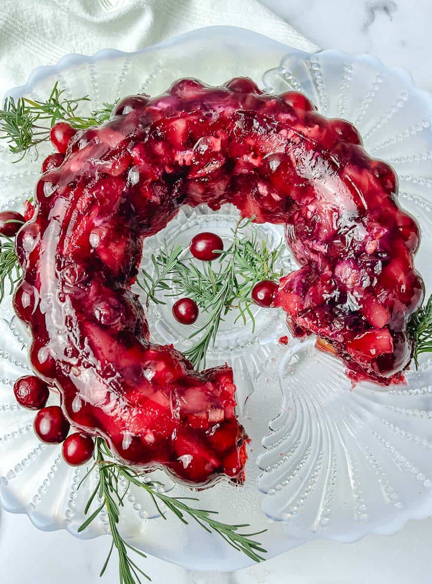 Cut cranberry pineapple jello salad mold on a glass plate with pineapple tidbits and cranberries showing, rosemary and fresh cranberries around it
