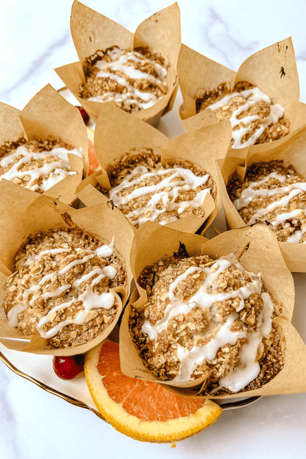 Easy cranberry orange muffins with oat streusel and white glaze drizzle in kraft tulip liners, served with an orange slice and fresh cranberries on a plate.