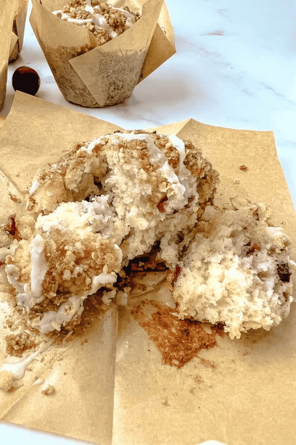 A cranberry orange muffin with oat streusel and glaze broken open on an unwrapped tulip liner, showing the soft, fluffy inside, with another muffin behind it.