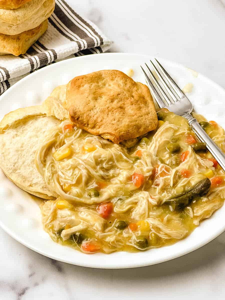 Crockpot chicken pot pie with creamy shredded chicken and vegetables on a white plate with a golden biscuit, a fork, and a striped towel