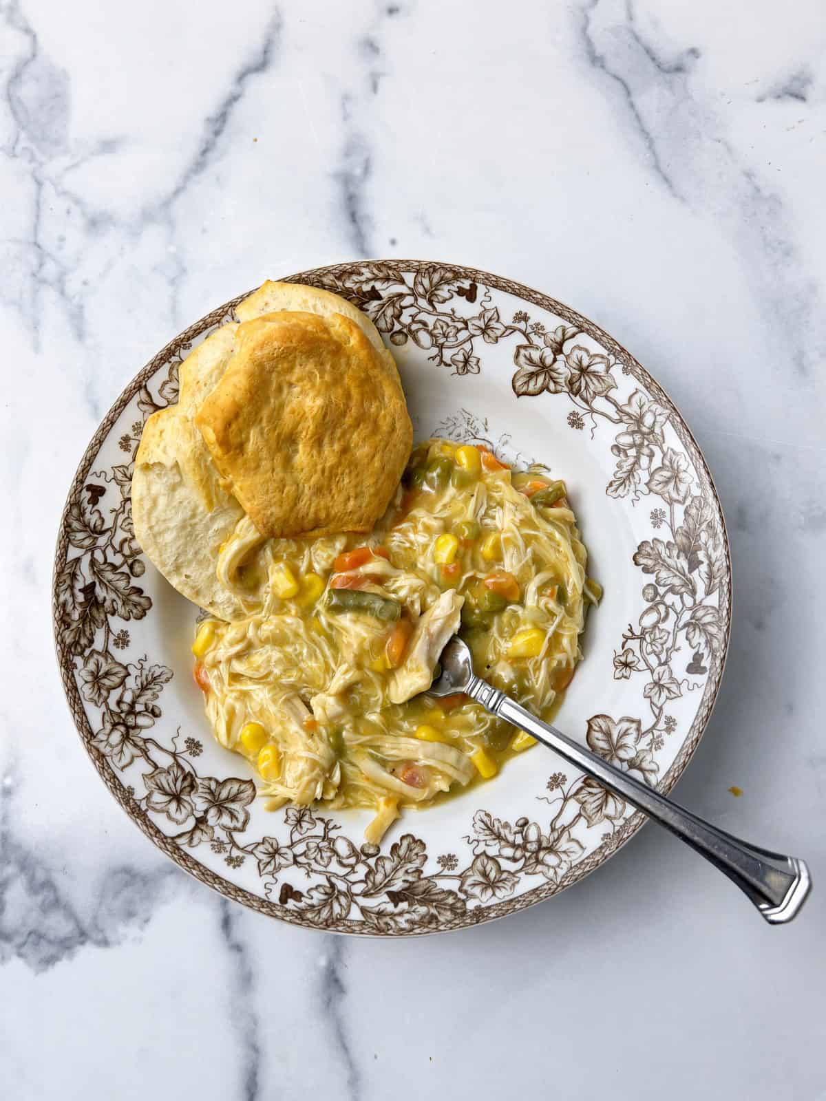 Crockpot chicken pot pie in a leaf patterned bowl with a baked biscuit and a spoon on white marble