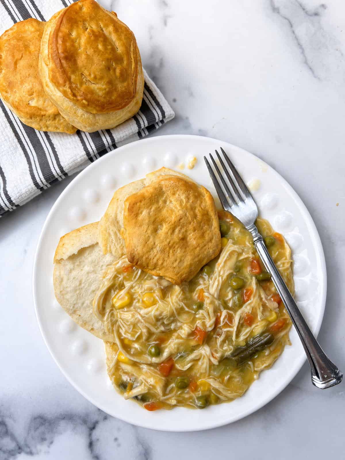 Crockpot chicken pot pie served on a white plate with a golden biscuit and a fork, next to two more biscuits on a striped towel