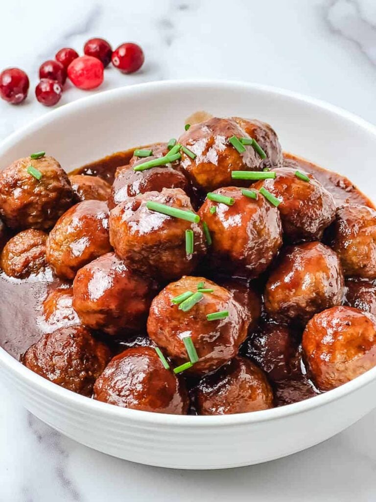Crockpot cranberry meatballs glazed in a glossy cranberry chili sauce in a white bowl, topped with chopped chives, with fresh cranberries on a white marble surface.