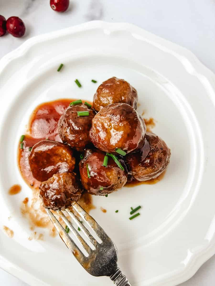 Close up of crockpot cranberry meatballs glazed in a sticky cranberry chili sauce on a white scalloped plate, topped with chopped chives, with fresh cranberries on the side for an easy holiday appetizer