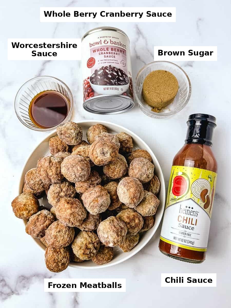 Ingredients for crockpot cranberry meatballs on a white surface, including a bowl of frozen meatballs, a can of whole cranberry sauce, a bottle of chili sauce, and small glass bowls of brown sugar and Worcestershire sauce