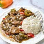 Crockpot pepper steak served over a mound of white rice on a white scalloped plate, with tender sirloin strips, thick sliced mushrooms and green and red bell peppers in a brown gravy, a silver fork on the side, a fresh green and orange pepper at the top, and a white marble background