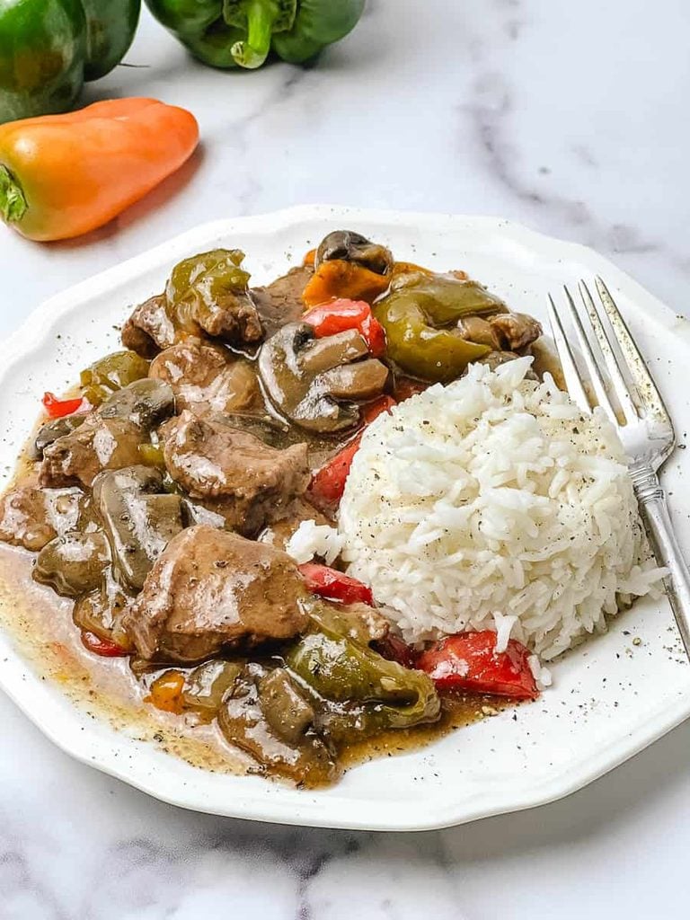 Crockpot pepper steak served over a mound of white rice on a white scalloped plate, with tender sirloin strips, thick sliced mushrooms and green and red bell peppers in a brown gravy, a silver fork on the side, a fresh green and orange pepper at the top, and a white marble background