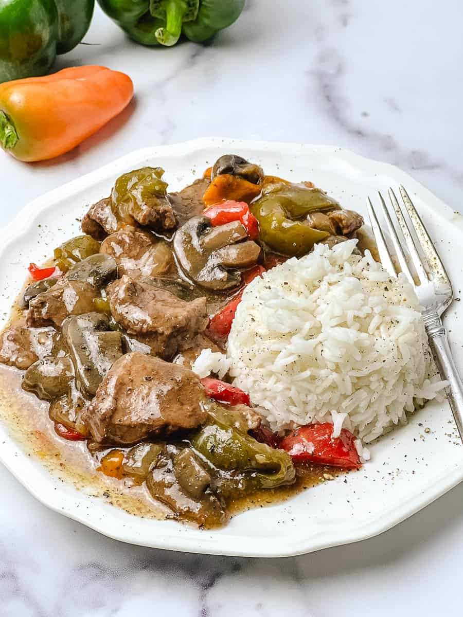 Crockpot pepper steak served over a mound of white rice on a white scalloped plate, with tender sirloin strips, thick sliced mushrooms and green and red bell peppers in a brown gravy, a silver fork on the side, a fresh green and orange pepper at the top, and a white marble background