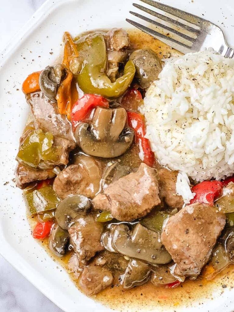 Close up of easy crockpot pepper steak with tender sirloin pieces, thick sliced mushrooms and green and red bell peppers in a brown gravy beside a mound of white rice on a white plate