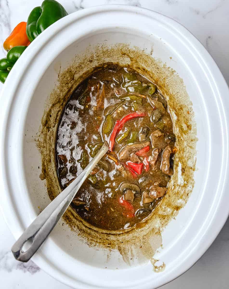 Easy crockpot pepper steak cooking in a white slow cooker with a spoon resting in the savory gravy, sliced green and red peppers, and a fresh green and orange pepper beside the pot
