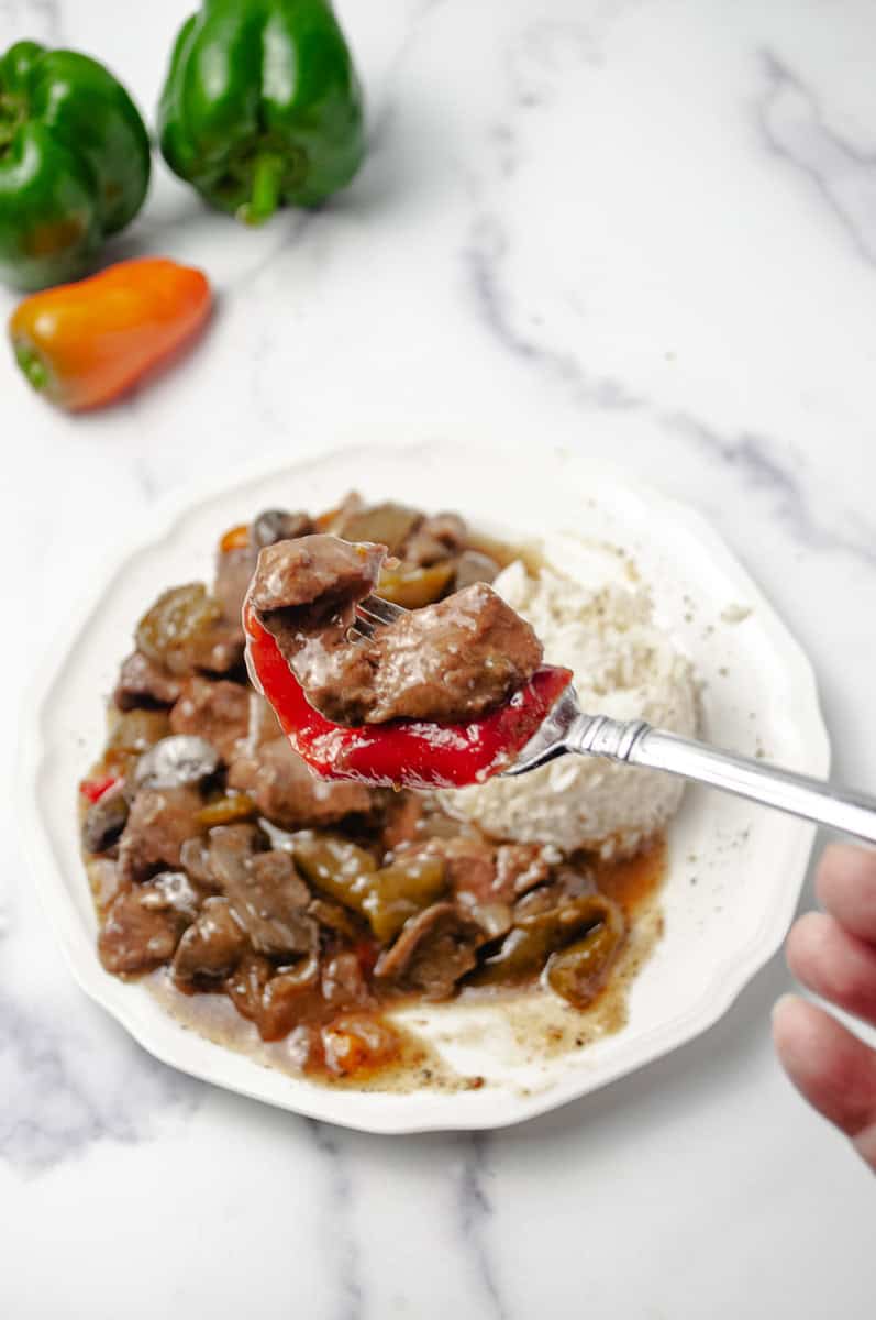 A forkful of tender pepper steak with a slice of red bell pepper lifted over a plate of crockpot pepper steak and white rice, with a fresh green and orange pepper on a white marble surface