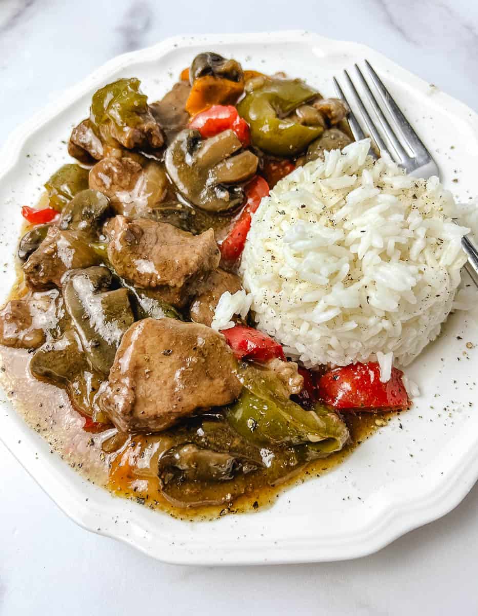Slow cooker pepper steak with tender sirloin strips, sliced mushrooms and green, red and orange bell peppers in a savory brown gravy next to a mound of white rice on a white plate