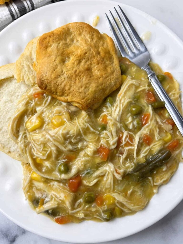 Close up of easy chicken pot pie in crockpot with shredded chicken, corn, carrots, and green beans on a white dotted plate with a golden biscuit and a fork