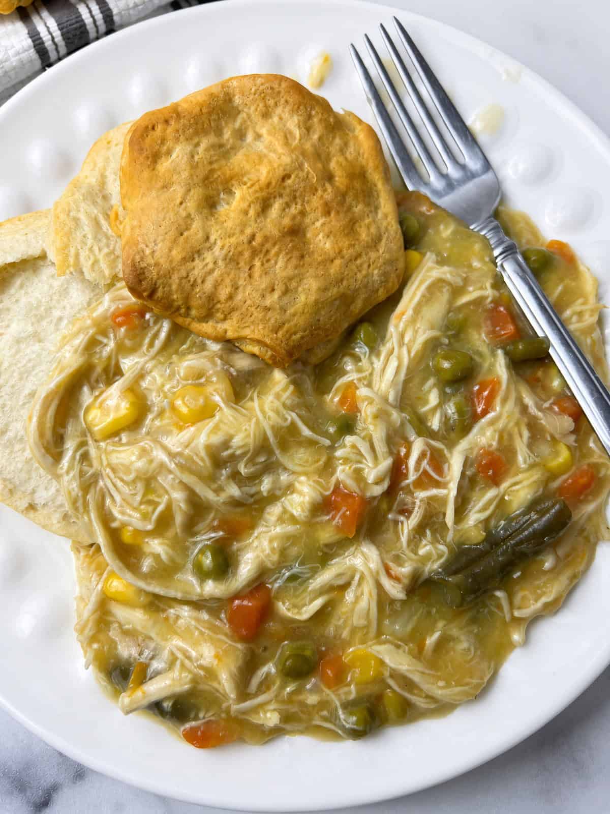 Close up of easy chicken pot pie in crockpot with shredded chicken, corn, carrots, and green beans on a white dotted plate with a golden biscuit and a fork