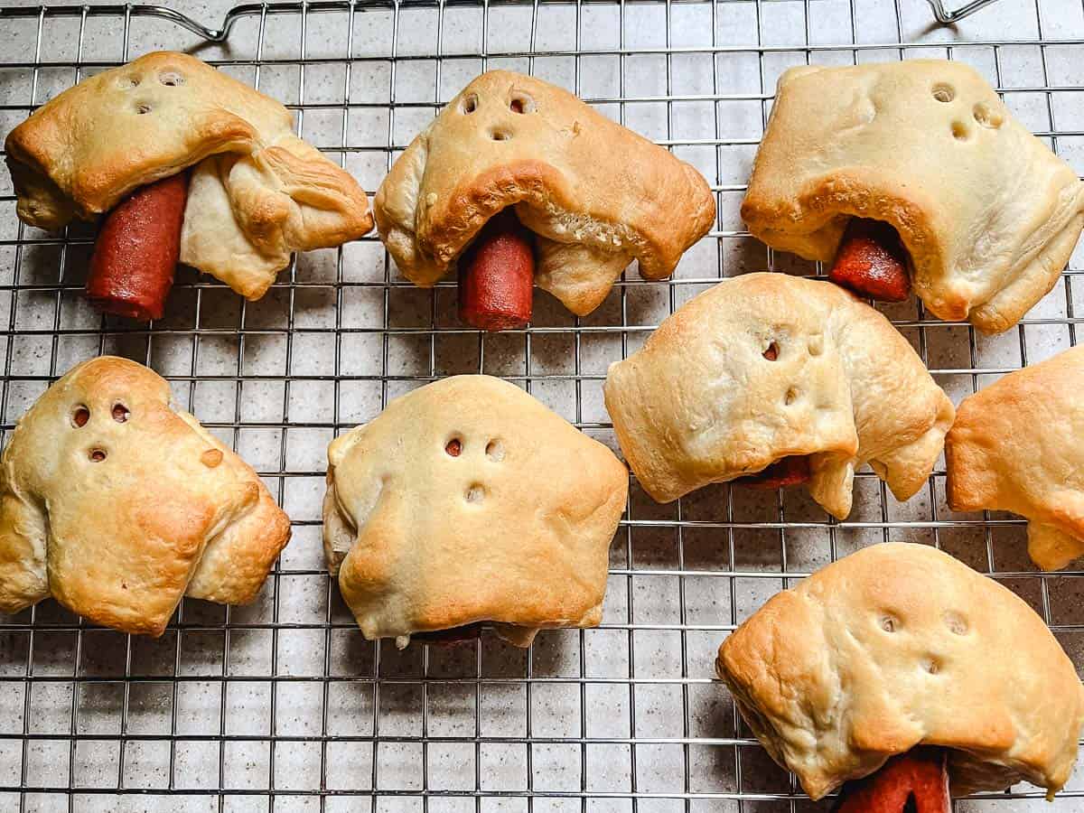 Golden baked Halloween hot dogs shaped like little ghosts, cooling on a wire rack.