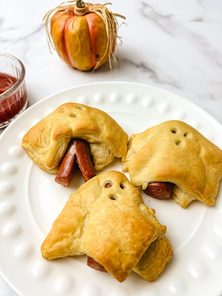 Ghost hot dogs made with crescent rolls on a white plate, with a small pumpkin and a dish of ketchup for dipping.
