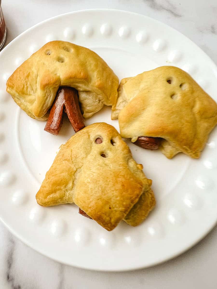 Three ghost pigs in a blanket on a white plate, an easy Halloween food idea for kids.