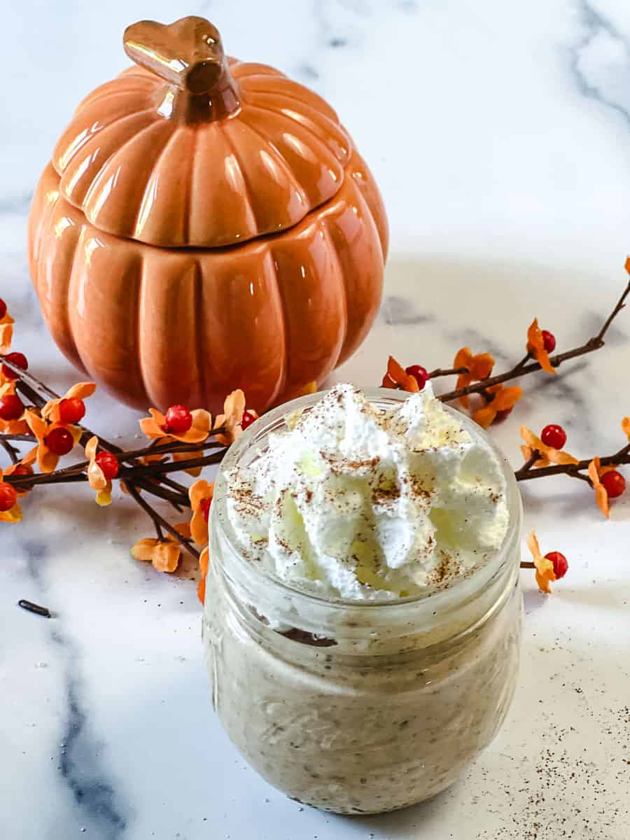 A jar of pumpkin chia pudding topped with whipped cream and cinnamon, set in front of an orange ceramic pumpkin with fall berry branches, a cozy no added sugar breakfast.
