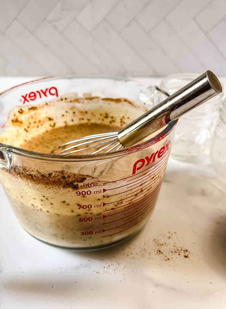 Pumpkin chia seed pudding mixture being whisked in a large glass measuring cup, with chia seeds and pumpkin pie spice swirled through the creamy Greek yogurt base.