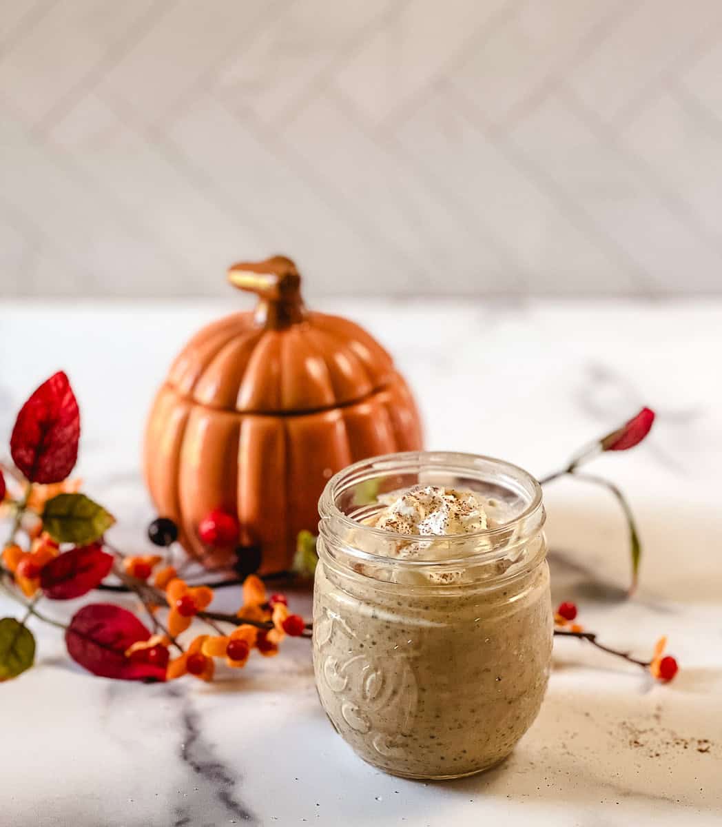 A glass jar of high protein pumpkin chia seed pudding with a small dollop of whipped cream, sitting on a marble counter with an orange ceramic pumpkin and red fall leaves behind it.