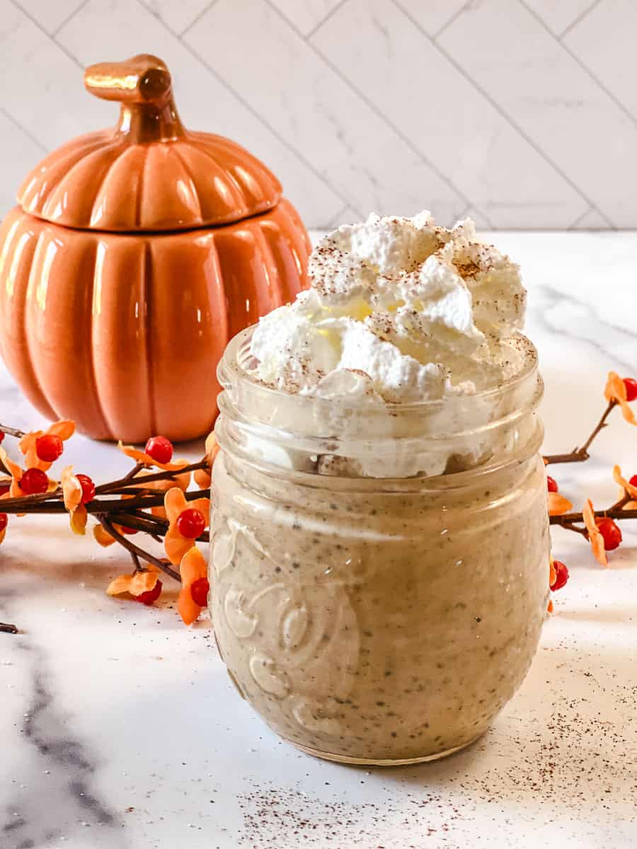Pumpkin chia seed pudding with Greek yogurt in a glass jar, topped with whipped cream and a sprinkle of cinnamon, next to an orange ceramic pumpkin and fall berry branches on a white marble counter.