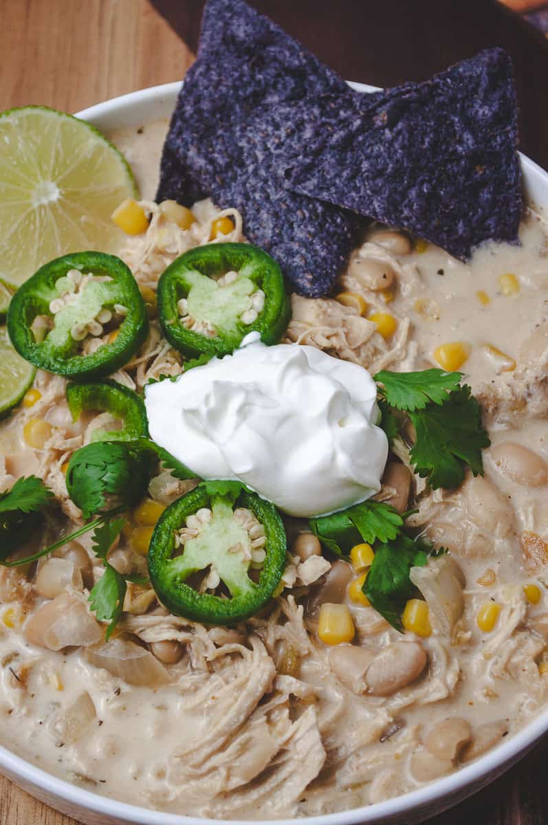 Close up of creamy white chicken chili made in the slow cooker, with shredded chicken, white beans, and corn in a white bowl, topped with sour cream, jalapeño slices, cilantro, a lime wedge, and two blue corn chips