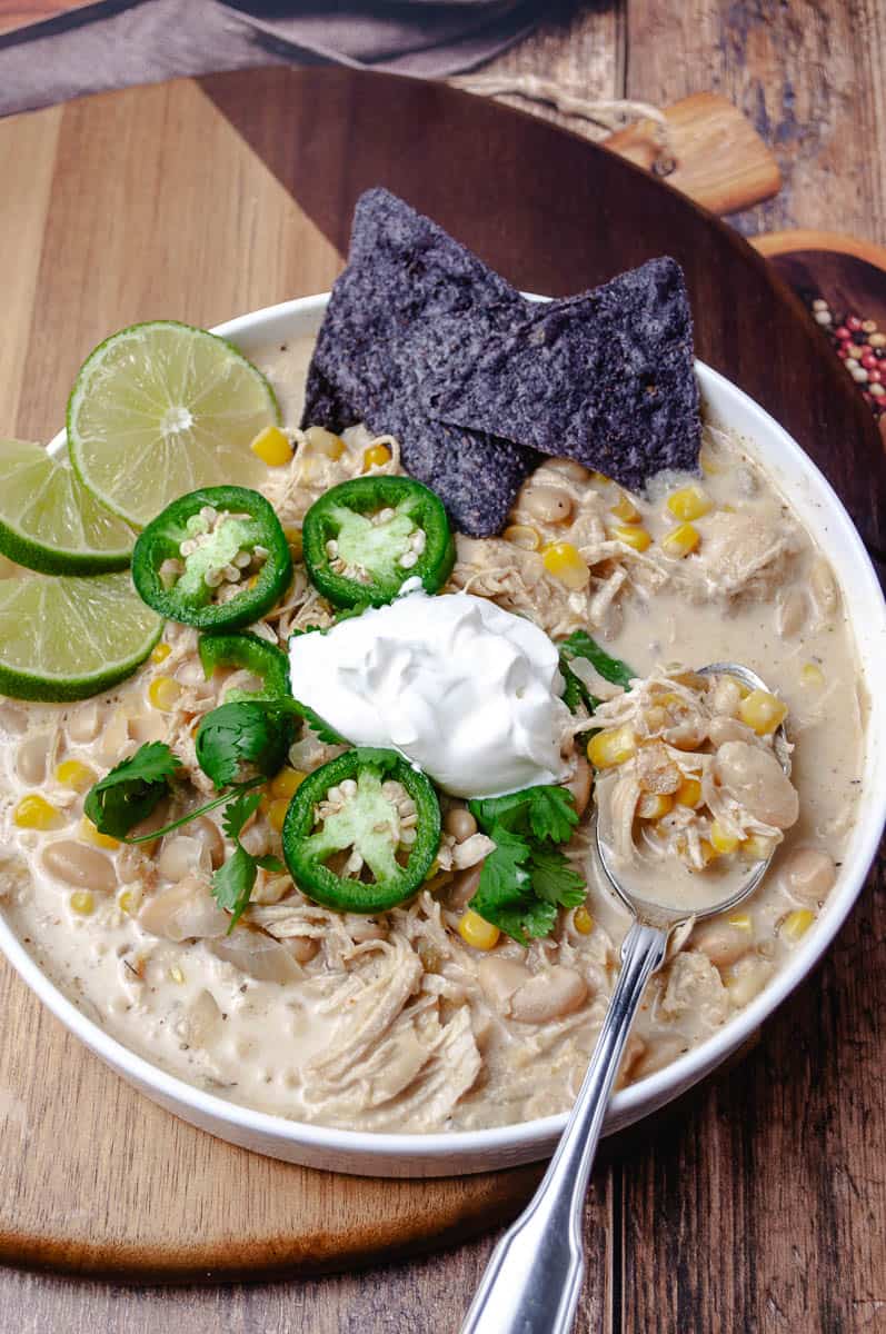 Creamy slow cooker white chicken chili in a white bowl with shredded chicken, white beans, and corn, topped with sour cream, jalapeño slices, cilantro, lime wedges, and blue corn chips, with a spoon lifting a bite on a round wooden board